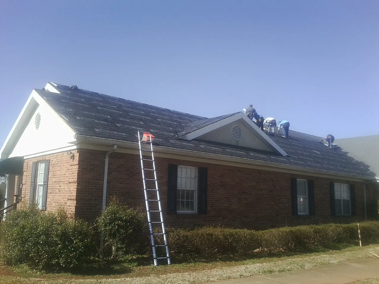 Crew tearing shingles off a brick ranch roof with a ladder set — Metal Roof Repair work by our crew in Henning